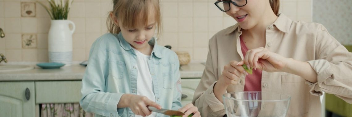 Adorable little girl is helping mother in kitchen cutting vegetables for salad and talking making dinner together. Healthy nutrition, lifestyle and people concept.